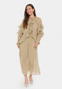 Beige, luftig klänning med volangdetaljer, långa ärmar och knytning vid halsen. Transparent material med ett lagerdesign och en midilängd.
