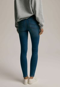 Chude dżinsy w ciemnoniebieskim denimu, z tylnymi kieszeniami z subtelnym przeszyciem, zestawione z szarym swetrem i białymi sneakersami.