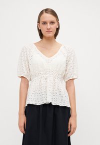 Weiße Bluse mit Puffärmeln und V-Ausschnitt, aus einem strukturierten, gemusterten Stoff mit taillierter Passform, kombiniert mit einem schwarzen Rock.