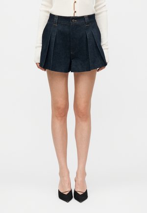 Kobieta w plisowanych ciemnych szortach z denimu, białym topie z długim rękawem i czarnych szpilkach z szpiczastym noskiem, stojąca na jednolitym białym tle.