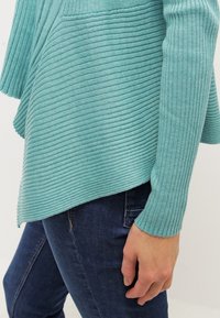 Top de punto acanalado en turquesa claro con dobladillo asimétrico y mangas largas, combinado con jeans azul oscuro, mostrando la posición de las manos.