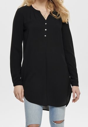 Sort langærmet tunika med V-hals, der har tre knapper ved halsudskæringen og en buet kant. Lavet af glat stof, båret over slidte jeans.