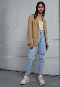 Beige oversized bleiseri yllä vaaleaa toppia, yhdistettyn�ä vaaleansinisiin korkeavyötäröisiin farkkuihin, joissa on reikä, ja beigen värisiin paksupohjaisiin lenkkareihin.