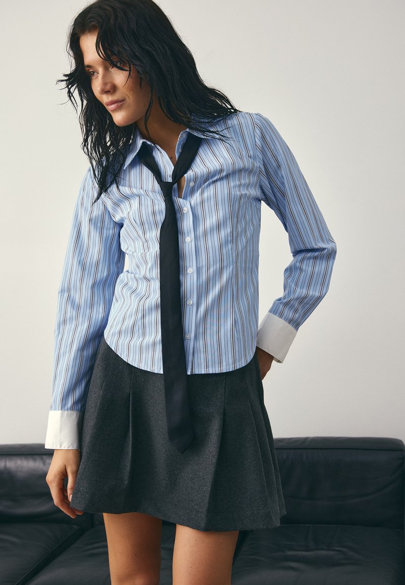 Moteris dėvi mėlyną dryžuotą marškinių su baltais rankogaliais, juodą kaklaraištį ir tamsiai pilką klostuotą sijoną, stovinti šalia juodos sofos.