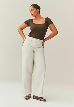 Woman with dark hair in a bun wearing a brown square-neck top, high-waisted white wide-leg pants, and snakeskin-patterned flats, posing with hands on hips.