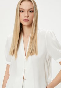 Blusa bianca a maniche corte con scollatura profonda a V e bottoni, realizzata in tessuto leggero. Caratterizzata da una texture liscia e una vestibilità morbida.