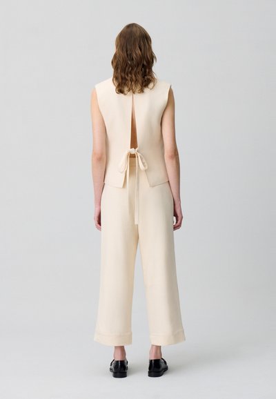 Combinaison sans manches beige avec un lien au dos et un design de dos ouvert. Présente des pantalons à jambes larges et des poignets. Texture lisse. Chaussures noires.