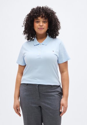 Donna con capelli ricci che indossa una polo azzurro chiaro a maniche corte e pantaloni grigi, in piedi davanti a uno sfondo bianco uniforme.
