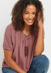 Bluzka z krótkim rękawem w kolorze mauve z gathered neckline, związywaniem i detalem w postaci frędzla. Gładki materiał o lekkim, luźnym kroju.