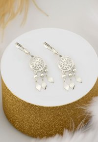 Silvriga örhängen med en cirkulär filigranmönster, med hängande silverbollar och diamantformade detaljer, visade på en vit piedestal.