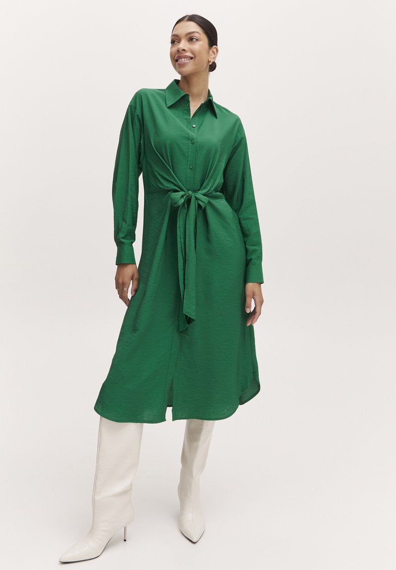 Langes grünes Hemdkleid mit durchgehender Knopfleiste und gebundenem Taillendetail, ausgestattet mit langen Ärmeln und einer glatten, glänzenden Textur. Kombiniert mit weißen kniehohen Stiefeln.