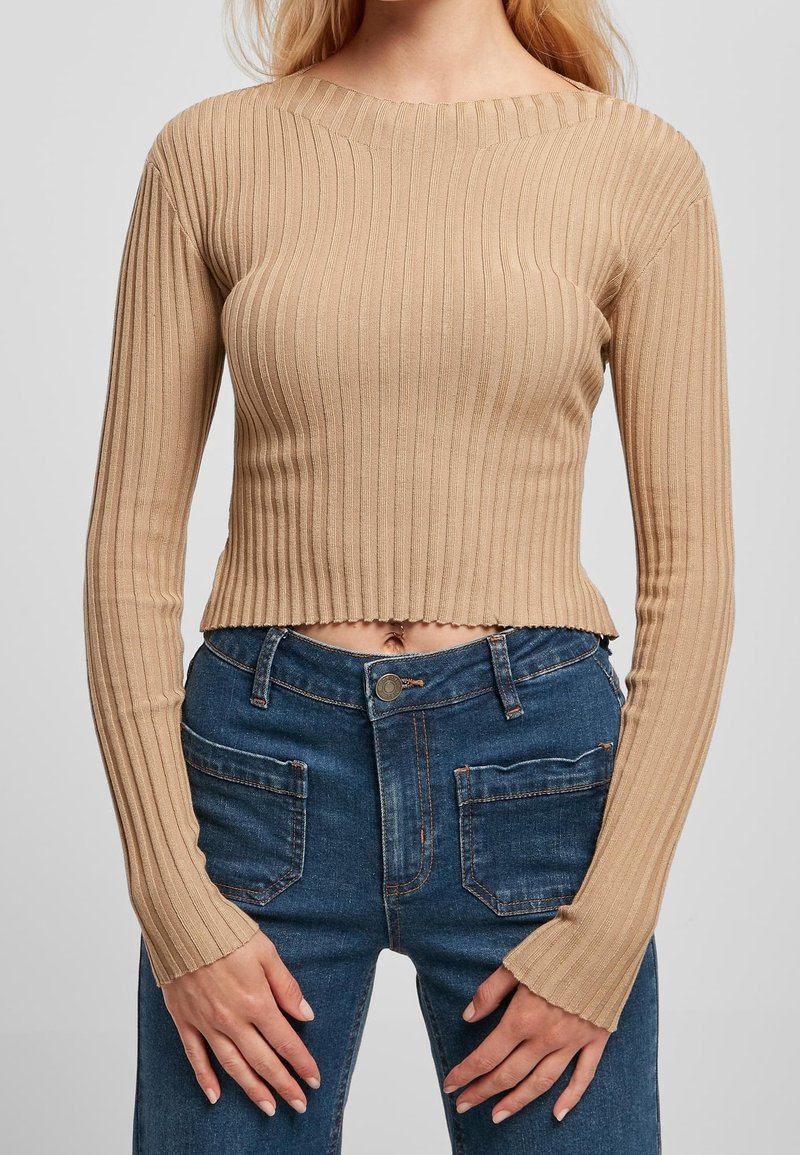 Femme portant un pull court côtelé beige à manches longues et un jean bleu taille haute avec poches avant, les mains reposant près des cuisses.