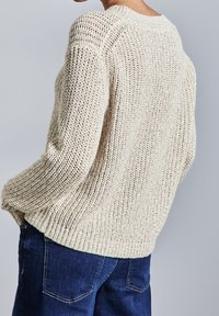 Pull en maille de couleur crème, présentant une coupe ample, un tissage ajouré texturé, des épaules tombantes et un ourlet côtelé. Associé à un jean bleu foncé.