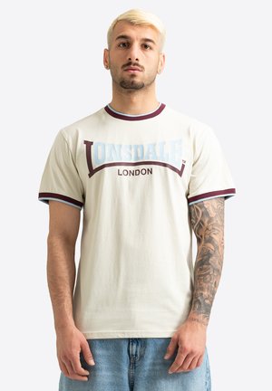 Mladi moški s kratkimi blond lasmi, oblečen krem Lonsdale London majico in modre kavbojke, ki kaže tetovažo z dolgimi rokavi na desni roki.