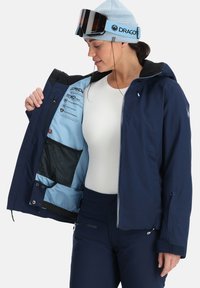 Marineblaue wasserdichte Skijacke mit schwarzer Innenauskleidung, Reißverschlusstaschen und einer inneren Netztasche, ausgestattet mit einer blauen Innenschicht und Logo-Akzenten.