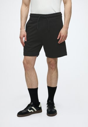Mand iført sorte shorts med snørre, sorte sokker og sorte sneakers med hvide detaljer, stående mod en ensfarvet hvid baggrund.