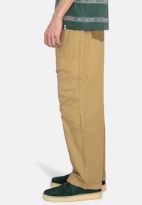 Vue de côté d'une personne portant un pantalon beige ample, des chaussures en suede vert foncé avec des semelles claires, et une chemise à motifs verts.