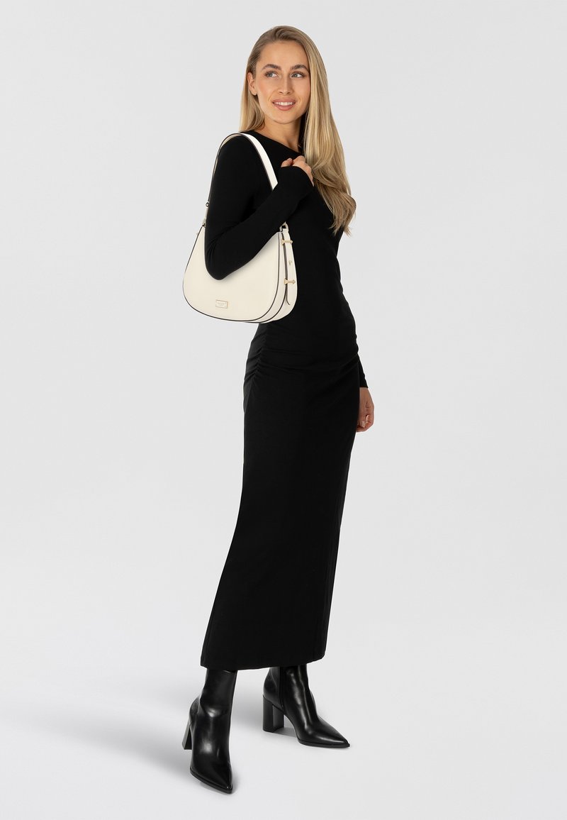 Zwarte lange mouw jurk en zwarte hooggekleurde laarzen tot aan de knie. Witte schoudertas met gouden accenten en zwarte bies. Model poseert met los haar.