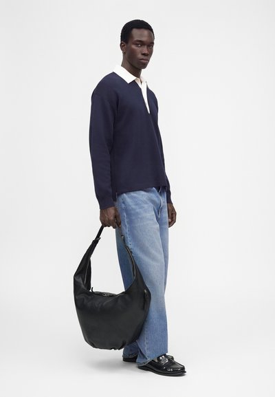 Un mannequin masculin se tient devant un fond blanc. Il porte une chemise bleu marine à col, un jean bleu clair et des chaussures noires, et tient un grand sac noir.