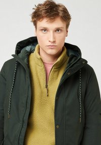 Jongeman met lichtbruin haar in een donkergroene winterjas met capuchon over een gele fleece met halflange rits.