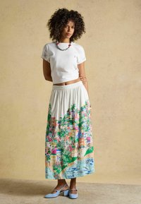 Femme aux cheveux bouclés portant un t-shirt court blanc, une longue jupe imprimée avec un paysage urbain coloré, des chaussures bleues à talons bas et un collier de perles.