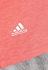 Červené sportovní tričko s texturovanou tkaninou, se bílým logem Adidas v rohu a stylovým střihem na spodním okraji.