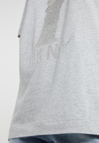 Grå bomulls-t-shirt med silver rhinestone-dekorationer som bildar bokstäverna "DKNY" och en grafisk design. Slät yta, rund hals.
