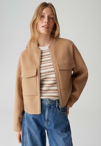 Giacca bomber color kaki realizzata in morbida lana, con zip frontale e due tasche sul petto. Maglione a righe in maglia sotto, con jeans di denim blu.