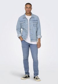 Svijetloplava traper jakna s dva džepa na prsima, nošena preko bijele majice, uparena sa svijetloplavim trapericama i crnim tenisicama.