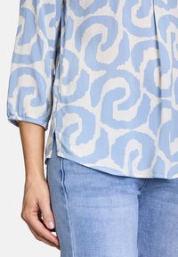 Blusa azzurra chiara con motivo bianco a spirali, maniche a tre quarti e spacco laterale; abbinata a jeans in denim azzurro chiaro.