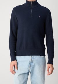 Marinblå zip-up tröja med texturerad stickad tyg, hög krage och ribbade ärmslut, kombinerad med ljusblå jeans. Subtil logotyp på bröstet.