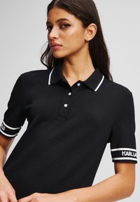 Mustapaitapaita, jossa on klassinen kaulukset, kolme nappia, valkoiset raidat hihoissa ja "KARL LAGERFELD" -logo hihansuissa.