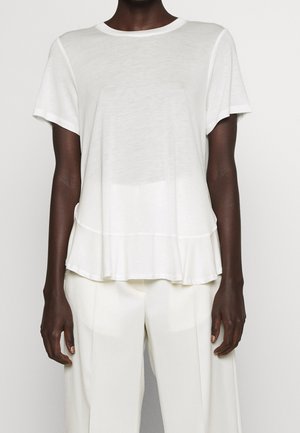 Henkilö, jolla on yllään lyhythihainen valkoinen paita, jossa on levenevä helma, ja väljät valkoiset housut, seisoo vaaleaa taustaa vasten.