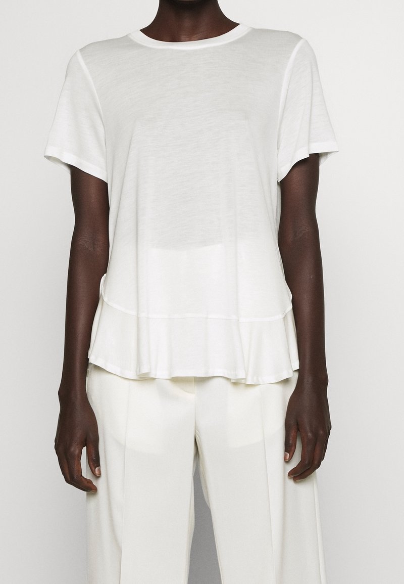 Personne portant un haut blanc à manches courtes avec un ourlet évasé et un pantalon blanc ample, se tenant devant un fond clair.