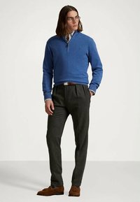 Μπλε πουλόβερ, μακρύ μανίκι, με φερμουάρ στον λαιμό και ραμμένα μανίκια, συνδυασμένο με γκρίζες παντελόνες και καφέ loafers. Το μοντέλο στέκεται σε απλό φόντο.