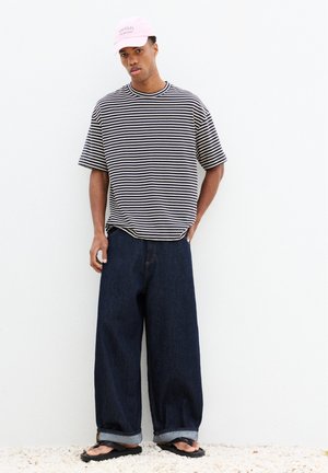 Jeune homme portant un t-shirt rayé noir et blanc, un jean large bleu foncé, des sandales noires et une casquette rose avec le texte « Amalfi Italian Coast ».
