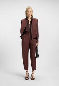 Gilet bordeaux sopra una blusa scura, abbinato a pantaloni sartoriali coordinati. Presenta linee eleganti, una zip e tacchi neri a strappo con una borsa.