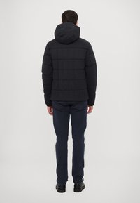 Dark quilted puffer jacket with a hood, featuring a rectangular pattern. Worn with dark pants and black shoes, viewed from the back.
