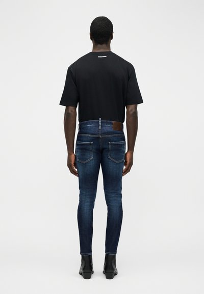 Homme aux cheveux courts portant un t-shirt noir, un jean skinny bleu foncé et des bottes noires, debout de dos sur un fond blanc uni.