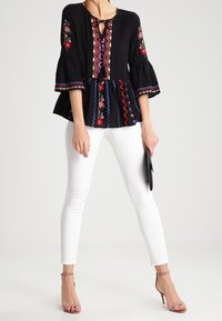 Top peplum nero con ricami floreali, scollatura a chiave e maniche a campana a 3/4. Abbinato a jeans skinny bianchi e tacchi rosa chiaro.