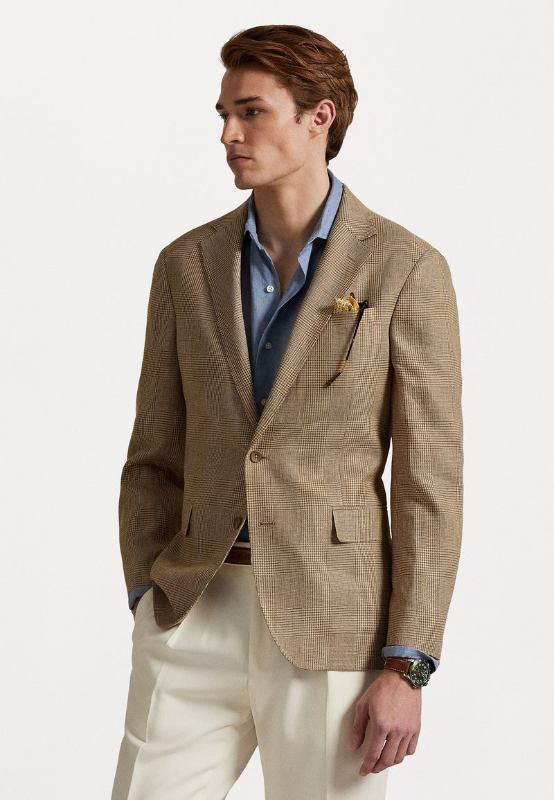 Bruine geruite blazer, op maat gemaakt, met één knoopsluiting, notch kent, gecombineerd met een lichtblauw shirt, beige broek en een bruin horloge.