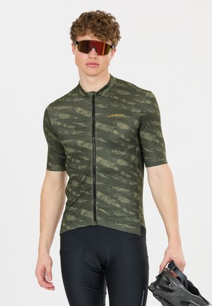 Jeune homme portant un maillot de cyclisme vert à motifs, un short de cyclisme noir, des lunettes de sport réfléchissantes, tenant un casque de vélo noir.