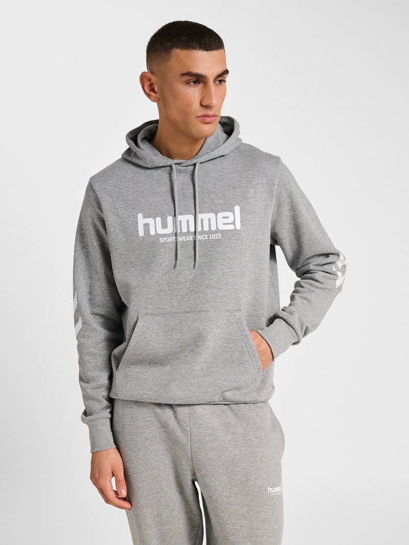 Man die een grijze hummel hoodie en bijpassende joggingbroek draagt, staat met één hand in de zak en kijkt opzij.