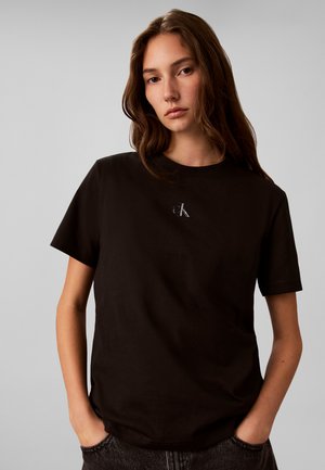 Jeune femme aux longs cheveux bruns portant un t-shirt noir Calvin Klein, debout les mains dans les poches devant un fond gris uni.