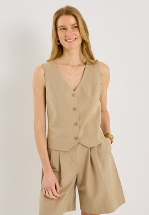 Femme souriante portant un gilet beige sans manches boutonné et un short plissé assorti, accessoirisée d'un collier doré, de boucles d’oreilles créoles et de bracelets.