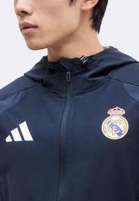 Νέος άνδρας φοράει μπλε μπουφάν Real Madrid με κουκούλα, κλειστό με φερμουάρ, με το λογότυπο της Adidas στο αριστερό στήθος και το έμβλημα του συλλόγου στο δεξί στήθος.