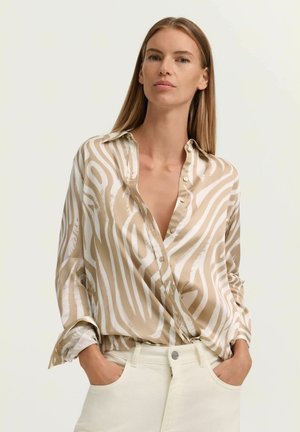 Woman with straight blonde hair wearing a beige and white zebra-print blouse and off-white pants, hands in pockets, neutral background.