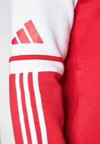 Röd och vit sweatshirt med röda detaljer, som har tre vita ränder och en röd Adidas-logotyp på vänster ärm. Mjukt tyg.