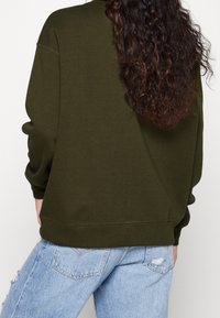 Sudadera verde oliva de corte holgado, con puños y bajo acanalados. Combinada con vaqueros azules desgastados y rotos, mostrando un estilo casual y en capas.