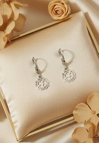 Silverfärgade blomformade hängande örhängen med utskuren design, fästa vid stängda klämmor, visade på en krämfärgad sidenkudde mot en mjuk, neutral bakgrund av tyg.